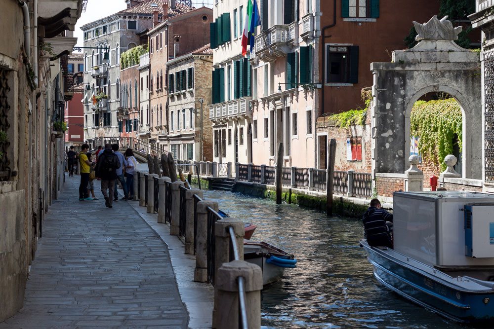 Небольшой канал в Венеции, вдаль уходят туристы, рядом проплывает лодка